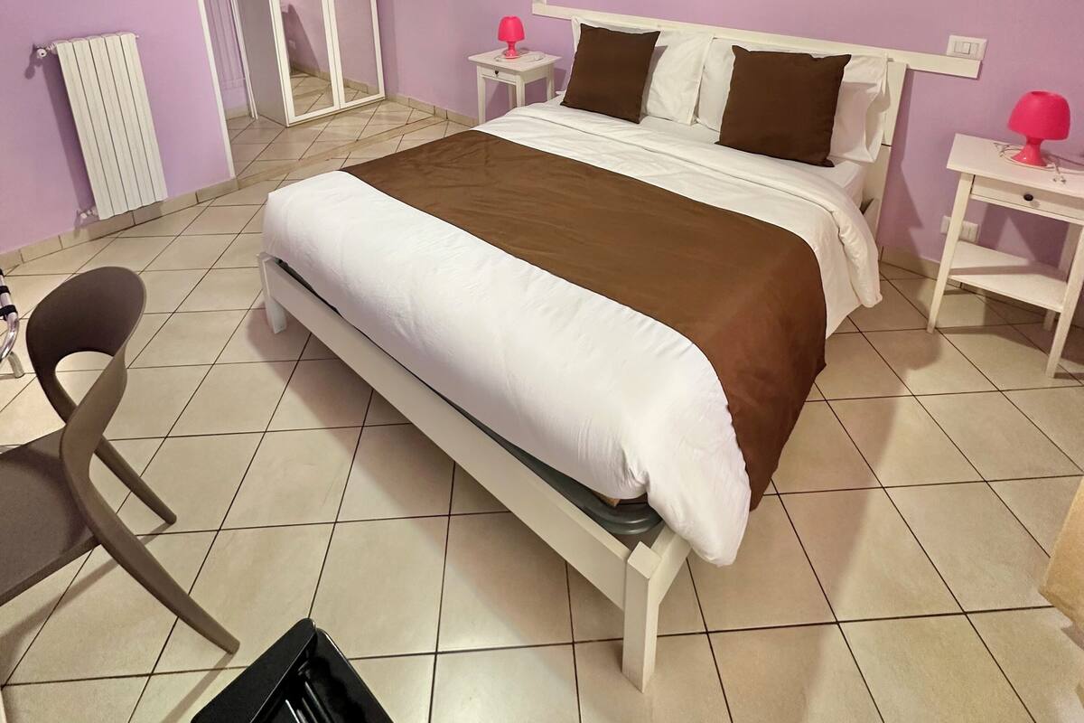 A bedroom with a bed, a chair, a desk, a wardrobe, and a nightstand with a lamp.
