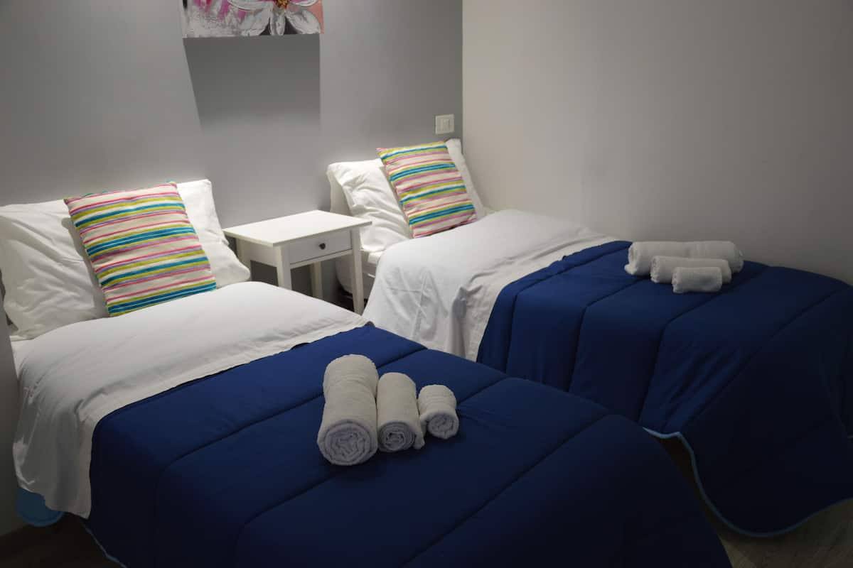 A hotel room with two beds, each with a blue bedspread and white pillows, one with a colorful striped pillow. There are rolled towels on the beds.