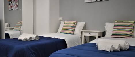 A hotel room with two beds, each with a blue bedspread and striped pillows. There are white nightstands with lamps and a small table between the beds.