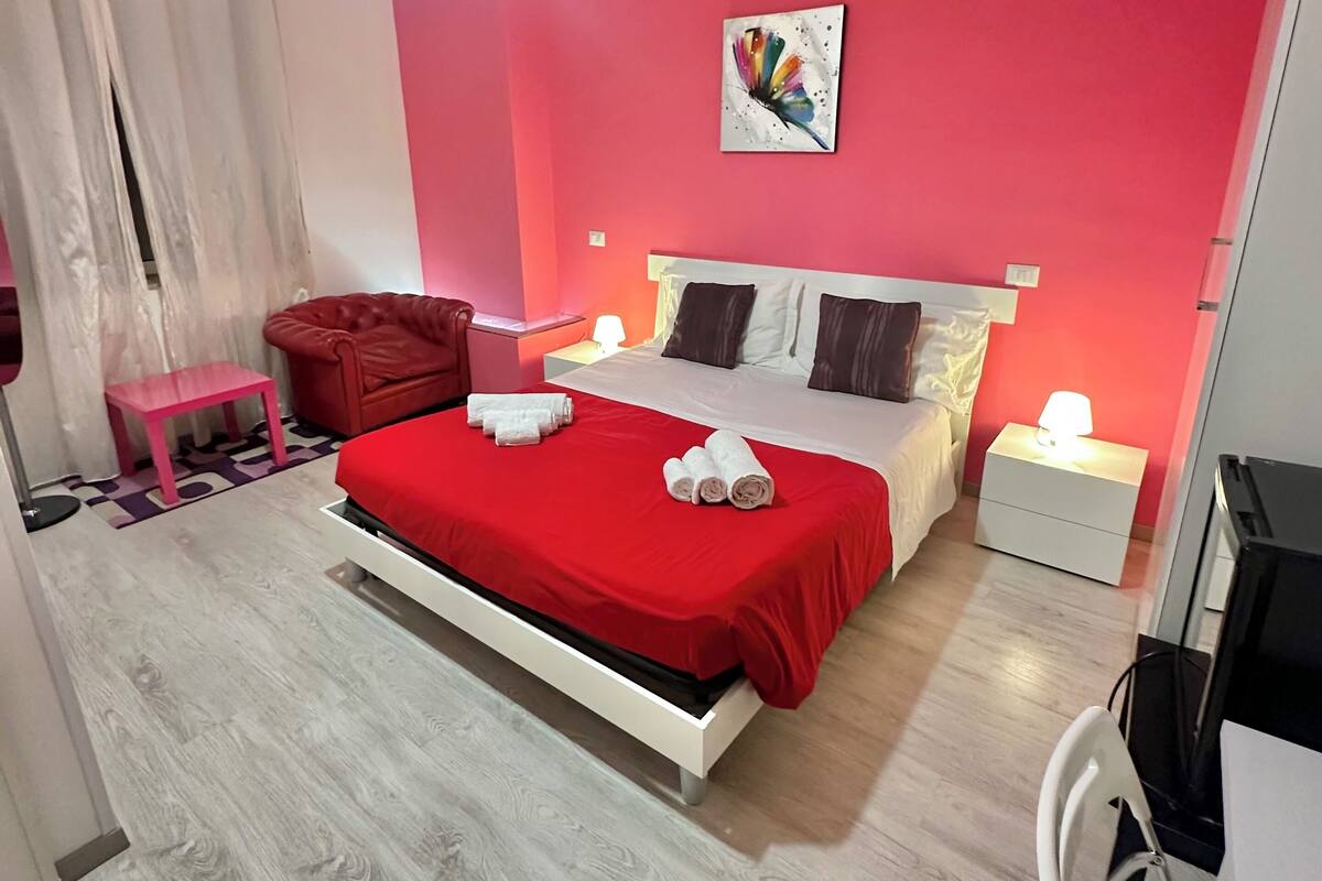A modern bedroom with a red accent wall, a bed with white bedding, a bedside table with a lamp, and a small pink stool.
