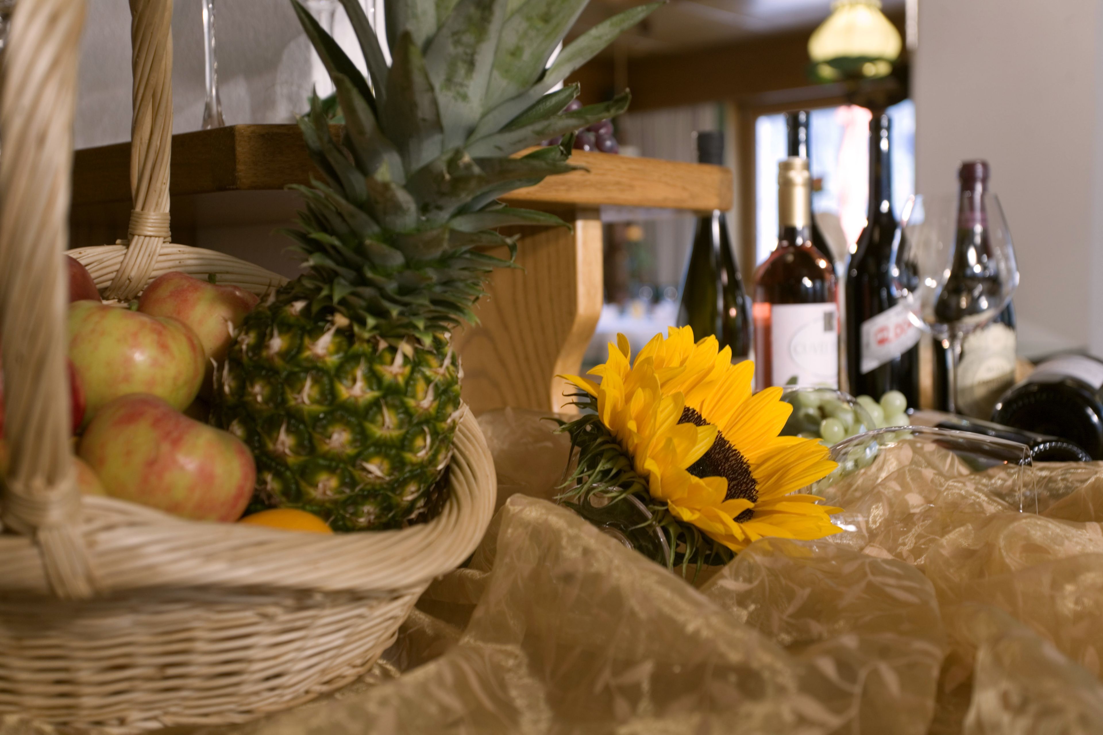A wicker basket with apples and a pineapple, a sunflower, and several wine bottles on a table.