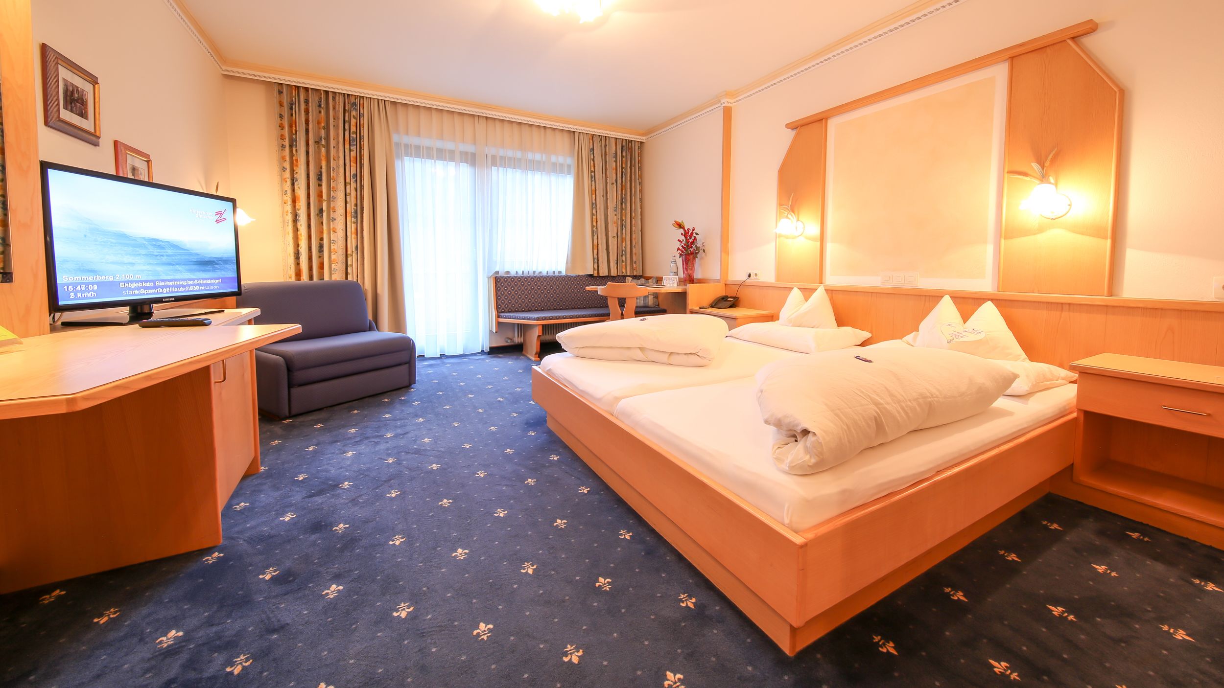 A hotel room with a bed, a desk, a sofa, and a TV displaying a flight information screen.