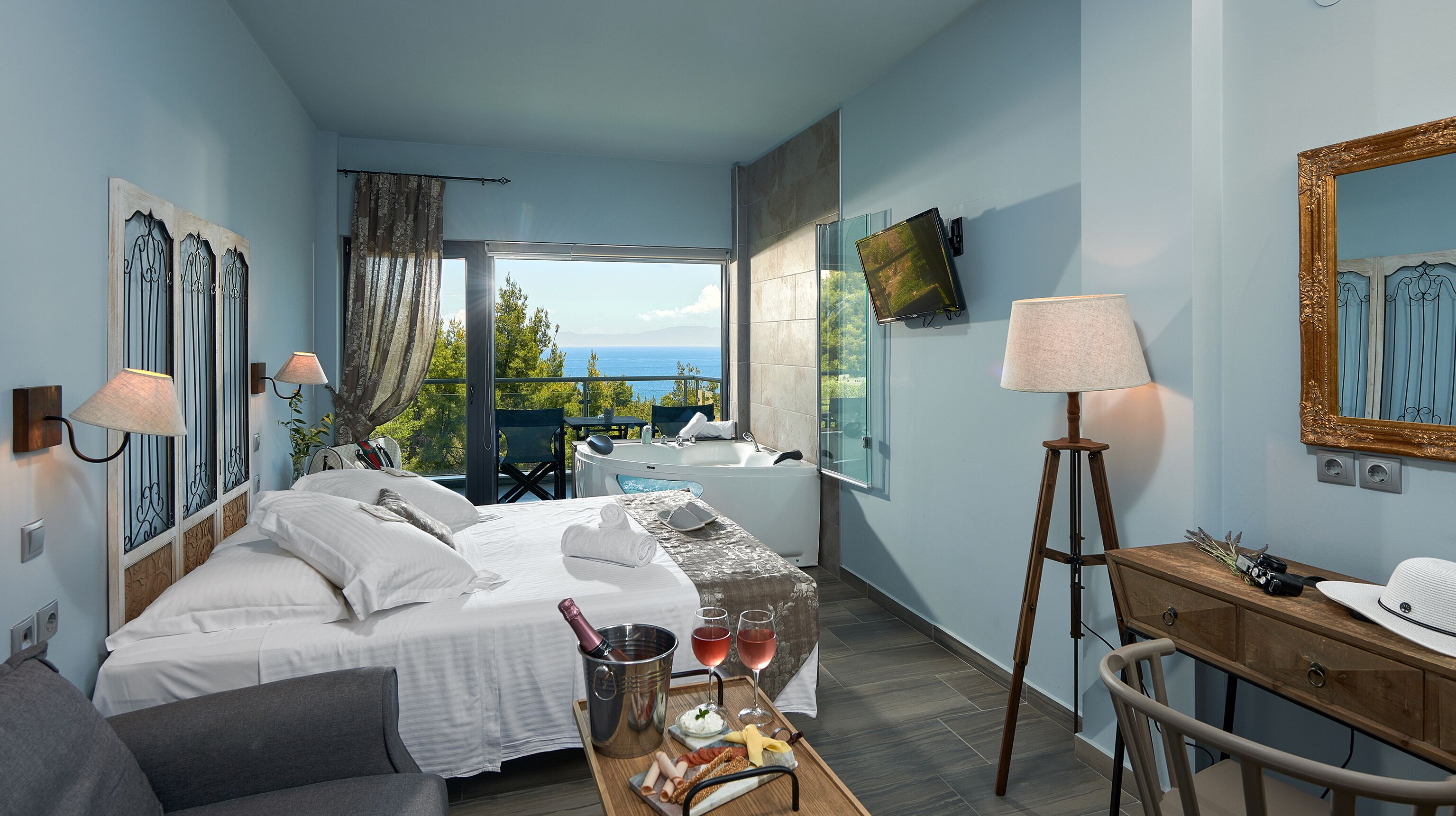 A hotel room with a bed, a desk, a chair, a table with wine glasses and a bottle, and a view of the sea.