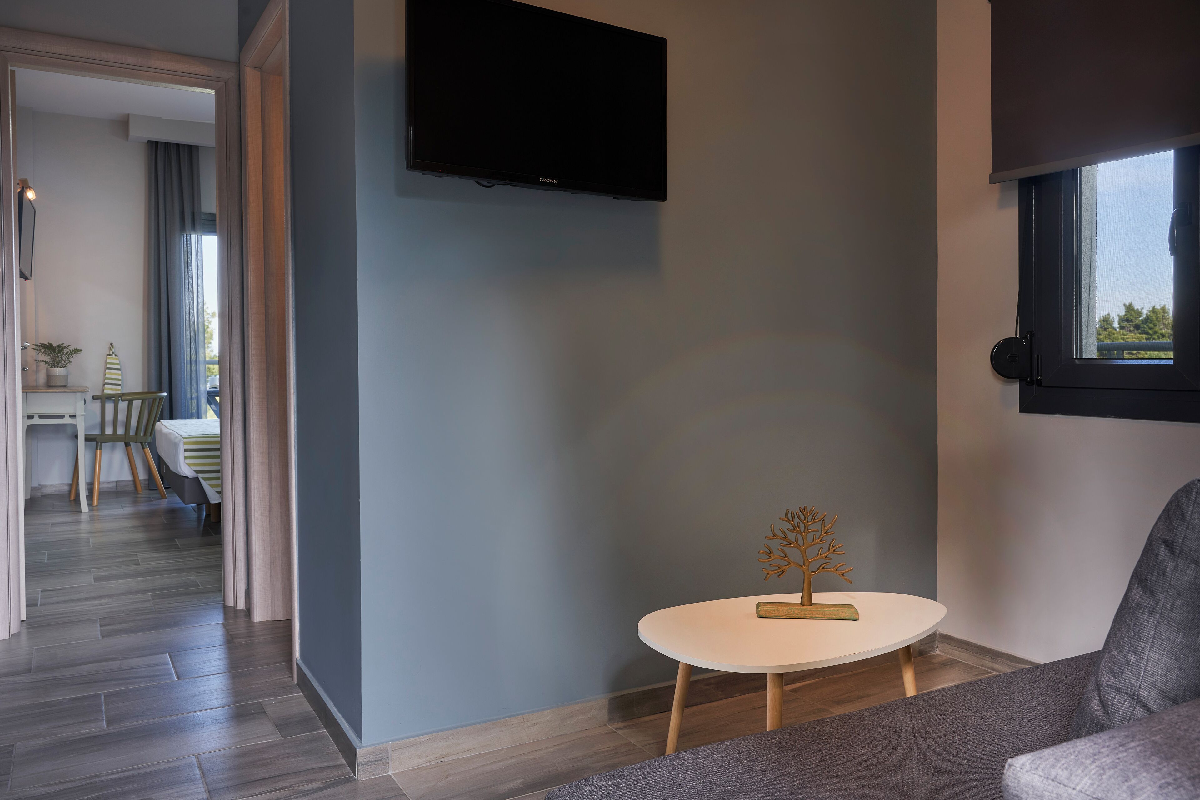 A modern living room with a flat-screen TV mounted on the wall, a round white table with a decorative tree, and a grey sofa.