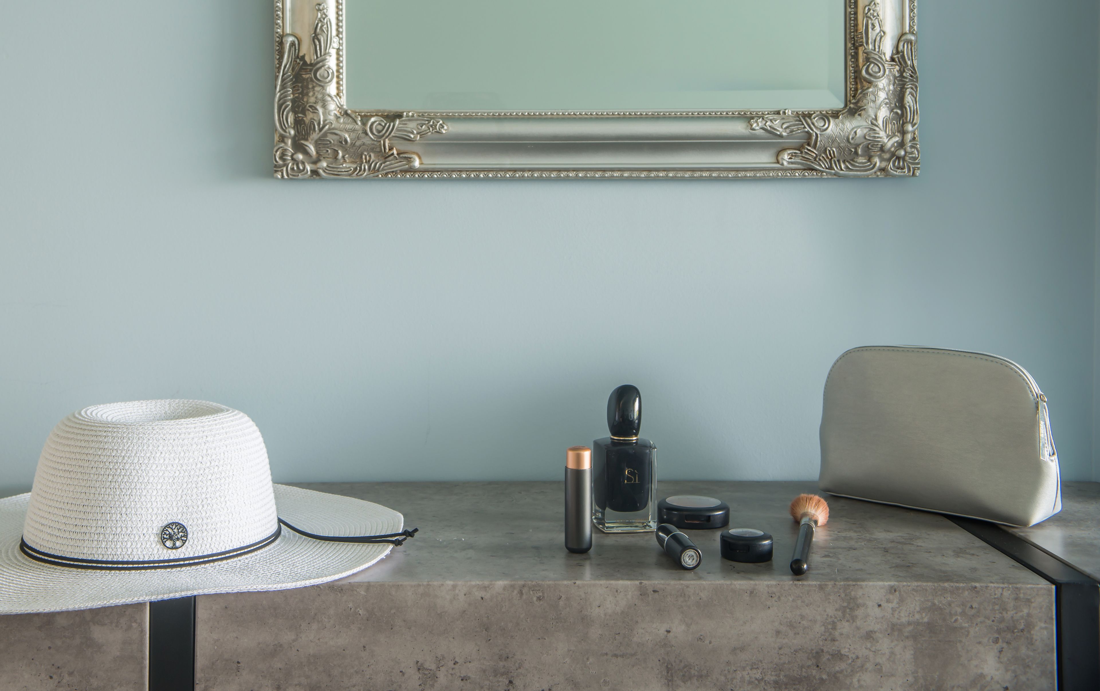 A room with a light blue wall, a framed mirror, a white hat, a black perfume bottle, a compact mirror, a brush, and a beige handbag on a concrete surface.