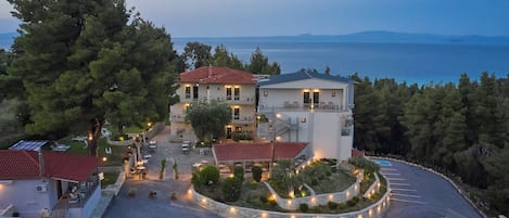Ένα παραθαλάσσιο ξενοδοχείο με κόκκινη κεραμοσκεπή, έναν περιποιημένο κήπο και θέα στη θάλασσα.