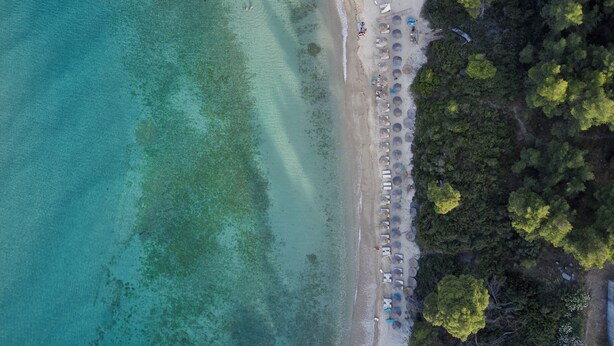 Vista aérea de una playa con agua turquesa cristalina, una orilla arenosa y una hilera de sombrillas y sillas de playa.