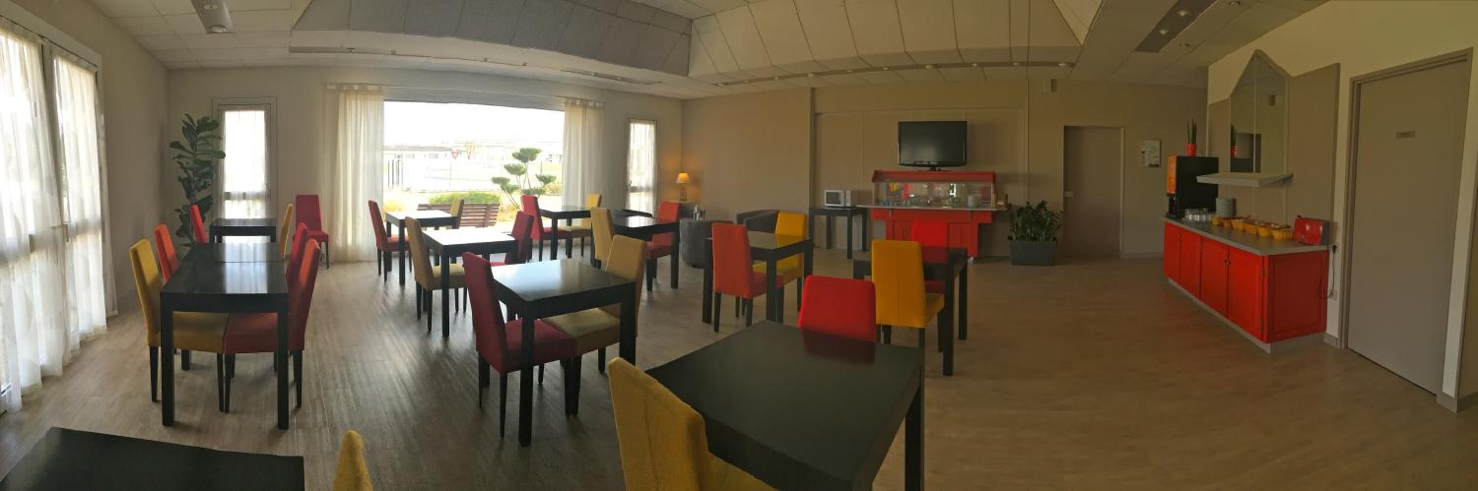 A spacious dining area with multiple tables and chairs, a kitchen area with red cabinets, and a television mounted on the wall.