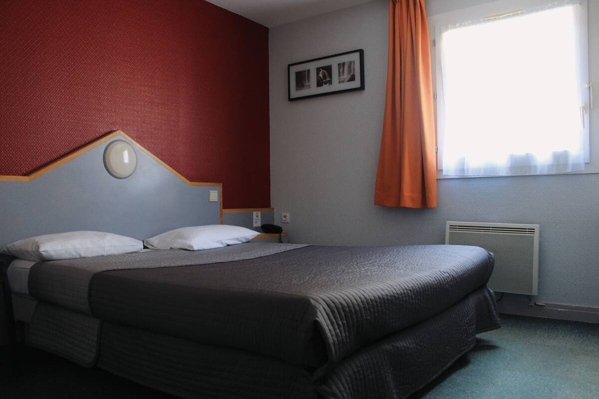 Ein Hotelzimmer mit Bett, Nachttisch, einem Bild an der Wand und einem Fenster mit Vorhängen.