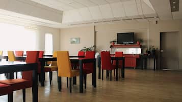 A dining area with multiple tables and chairs, a television mounted on the wall, and potted plants.