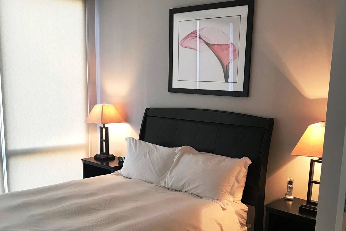 Un lit bien fait, avec du linge de lit blanc, une tête de lit sombre et deux lampes de chevet sur des tables de nuit. Un tableau encadré est accroché au mur au-dessus du lit.