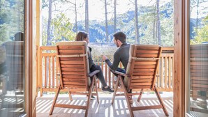 Dos personas sentadas en sillas de madera, frente a frente, en una terraza de madera con un muro de cristal, con vistas a una zona boscosa.