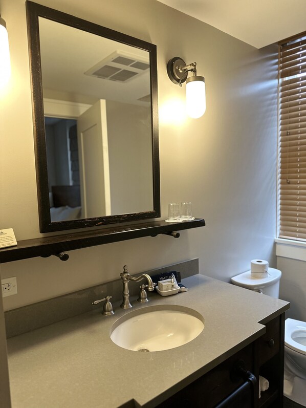Une salle de bain avec un grand miroir, un lavabo, un meuble-vasque, un porte-serviettes, des toilettes et une fenêtre avec des stores.