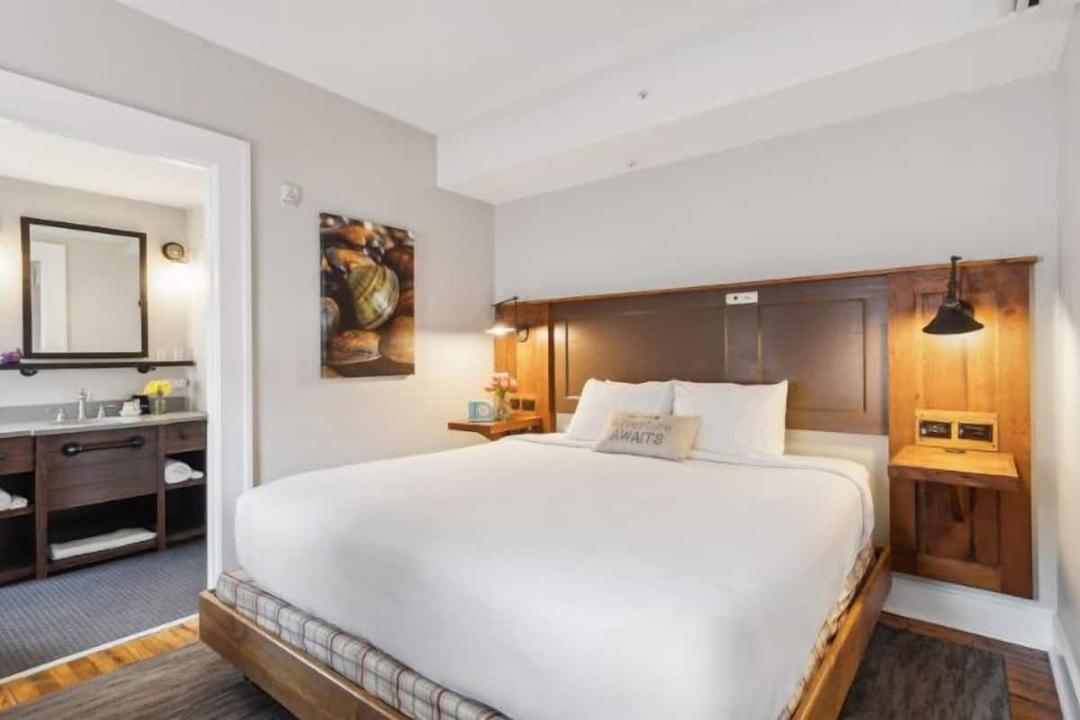 A hotel room with a large bed, a wooden headboard, a nightstand with a lamp, a mirror, and a painting on the wall.