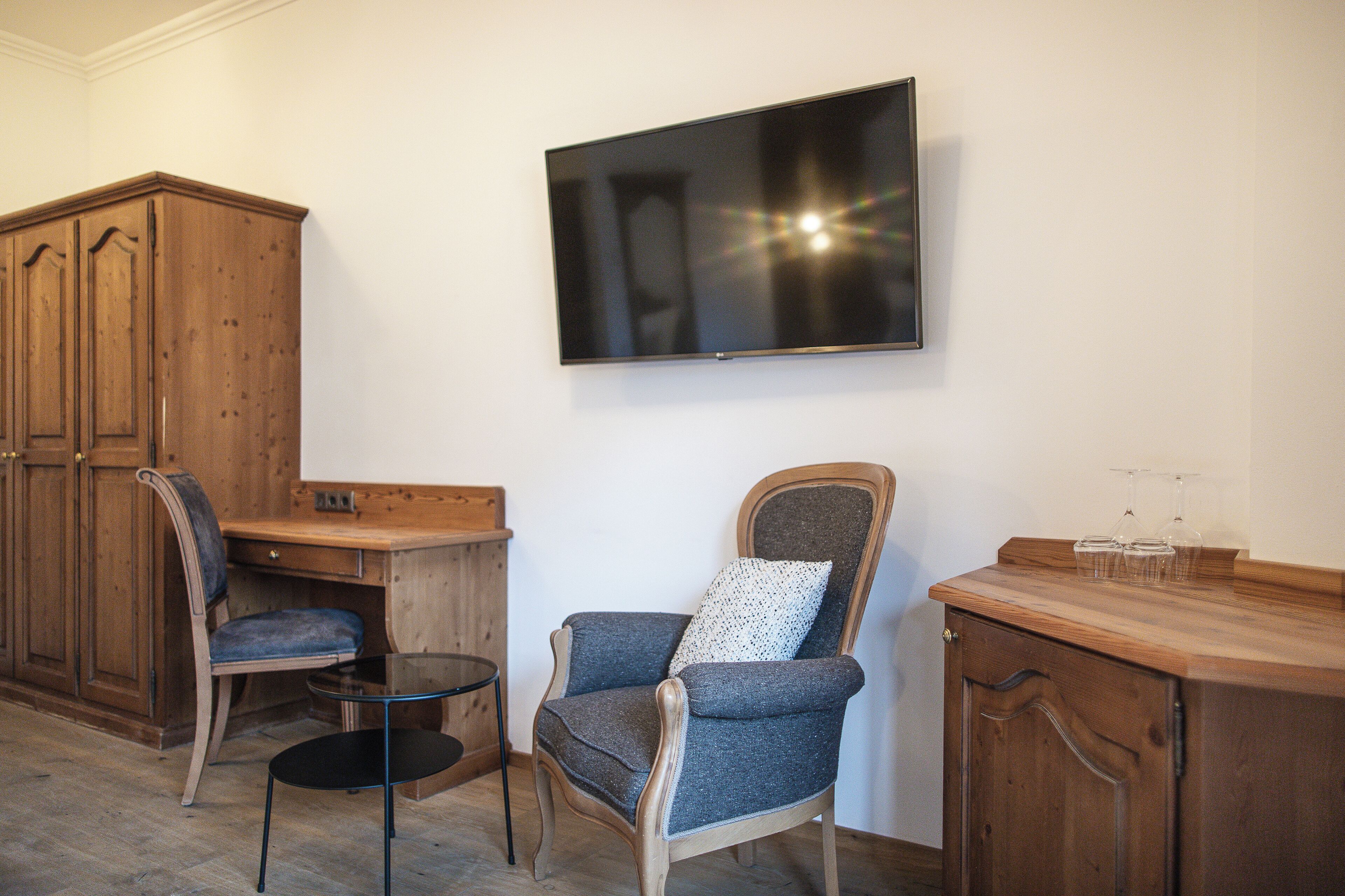 A room with a wooden cabinet, a desk, a chair, a television mounted on the wall, and a small table with glasses.