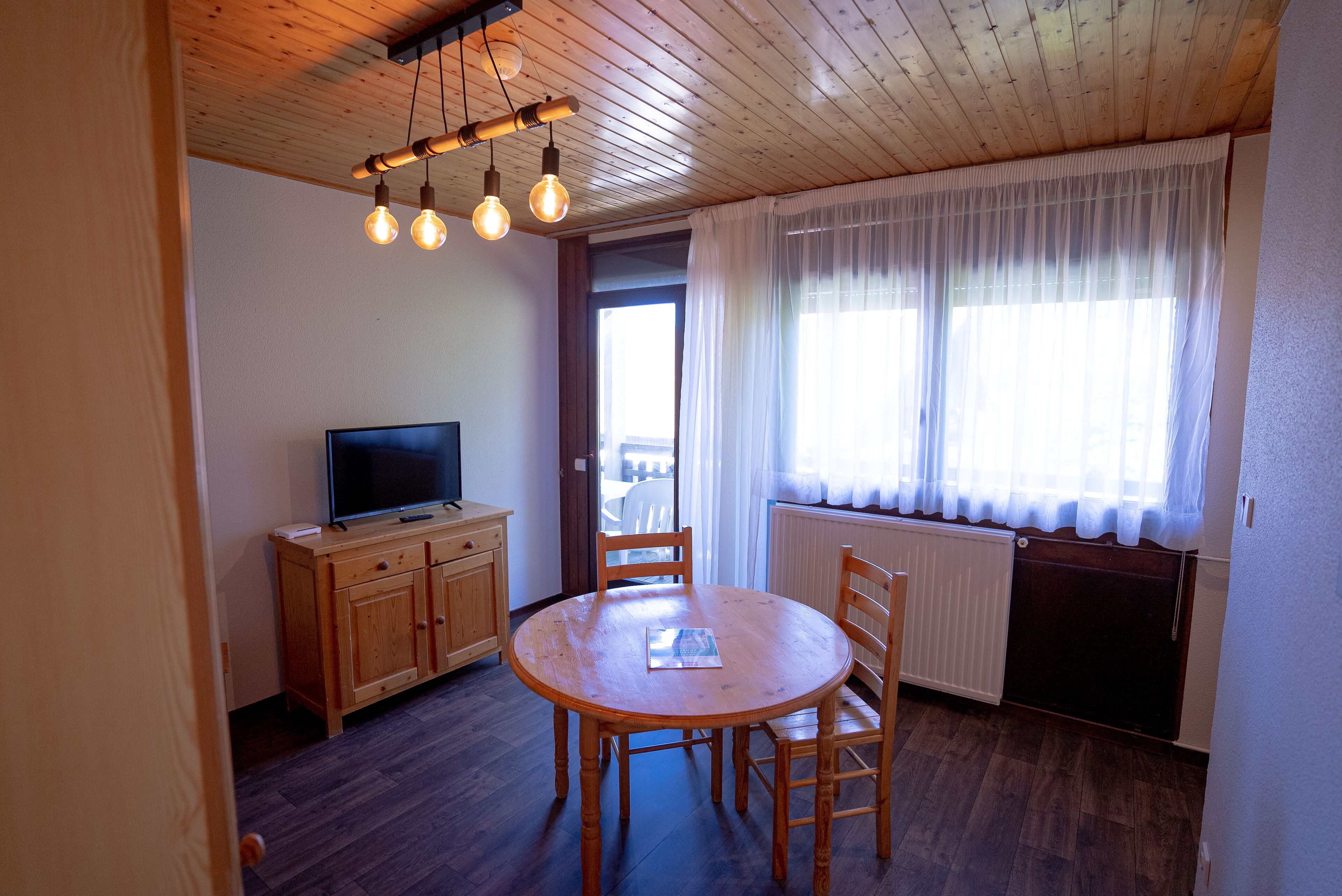 Un coin repas avec une table et des chaises en bois, un téléviseur posé sur un meuble en bois, et une fenêtre ornée de rideaux légers.