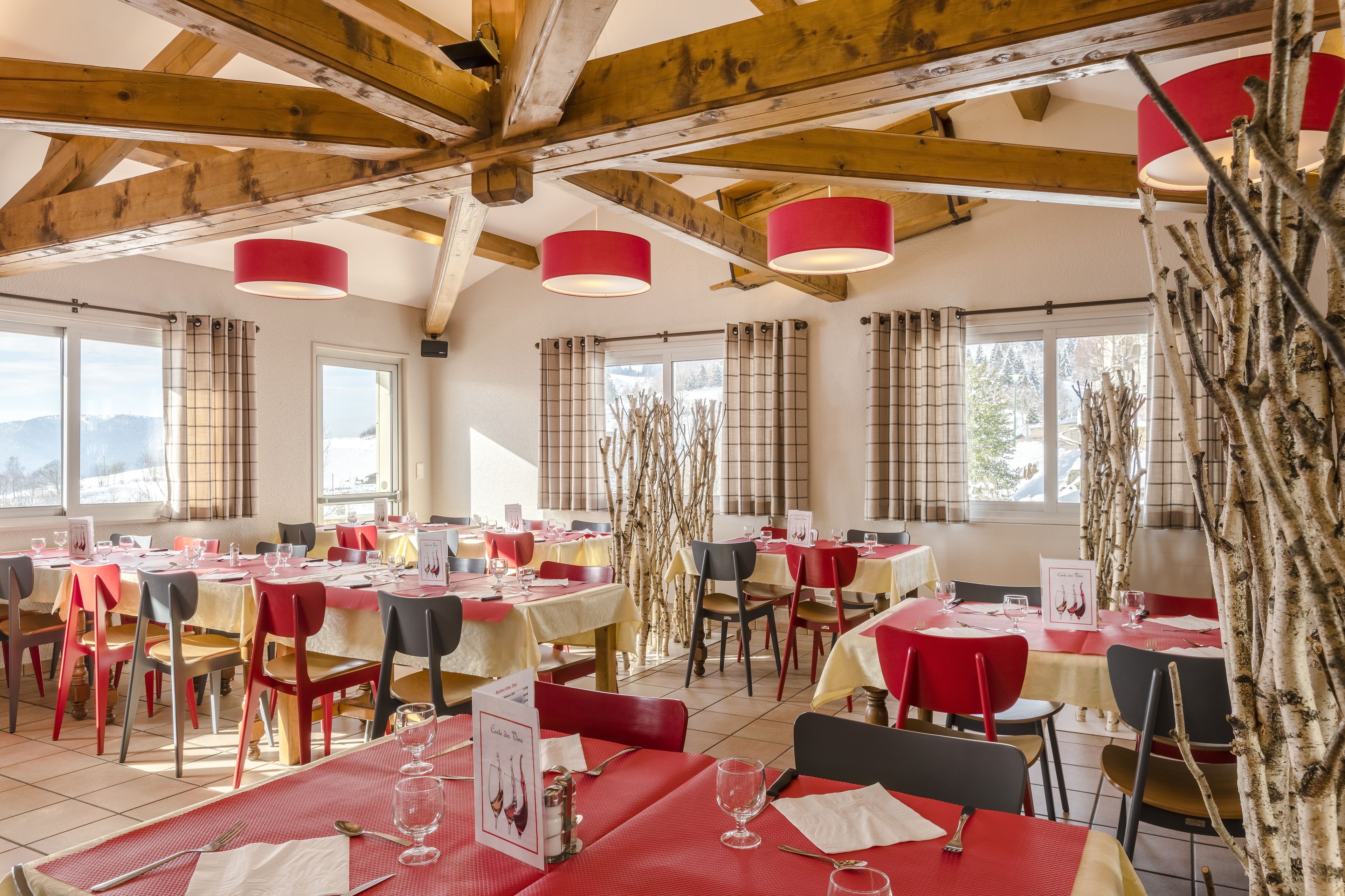 En restaurant med flere borde og stole, træbjælker i loftet og store vinduer med gardiner.