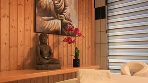 Un cuarto de spa con pared de madera, una estatua de Buda, una planta en maceta con flores rosadas, una toalla blanca y una camilla de masaje.