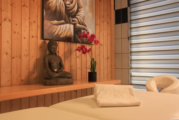 Un cuarto de spa con pared de madera, una estatua de Buda, una planta en maceta con flores rosadas, una toalla blanca y una camilla de masaje.