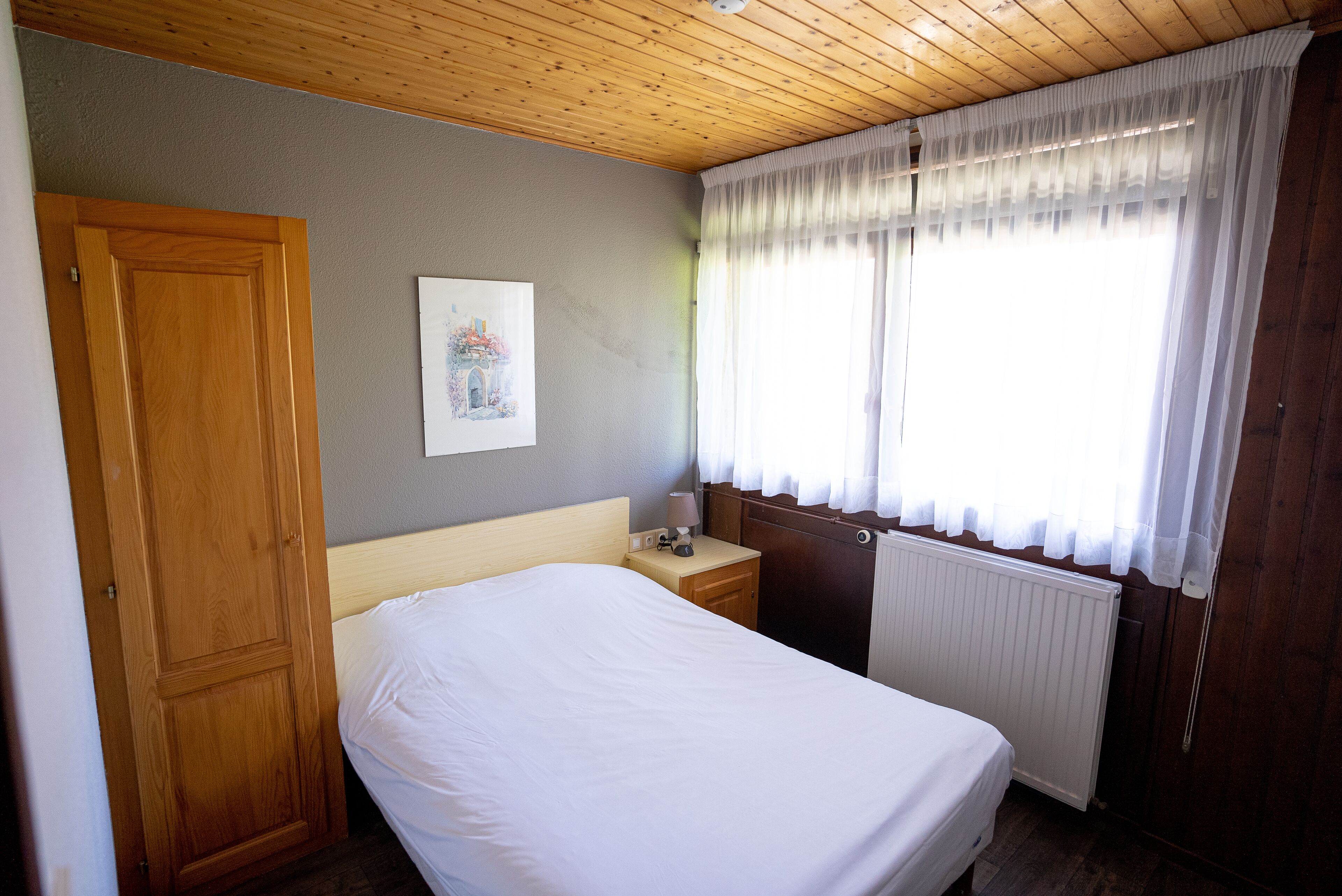 Een slaapkamer met een houten deur, een nachtkastje met een lamp, een radiator en een raam met dunne gordijnen.