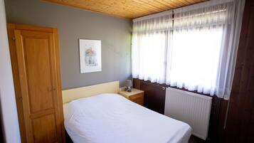 Een slaapkamer met een houten deur, een nachtkastje met een lamp, een radiator en een raam met dunne gordijnen.