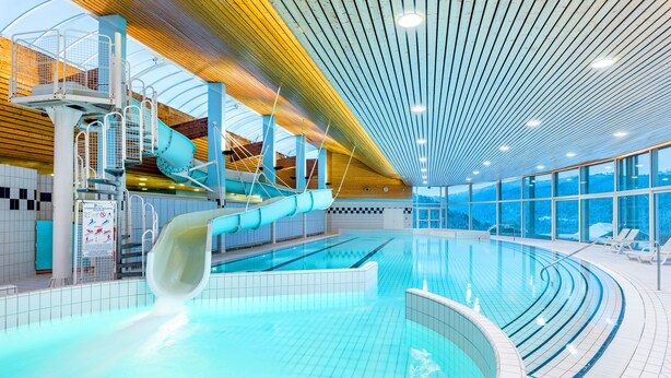Una gran piscina interior con tobogán, plataforma de salto y vistas al exterior a través de amplias ventanas.