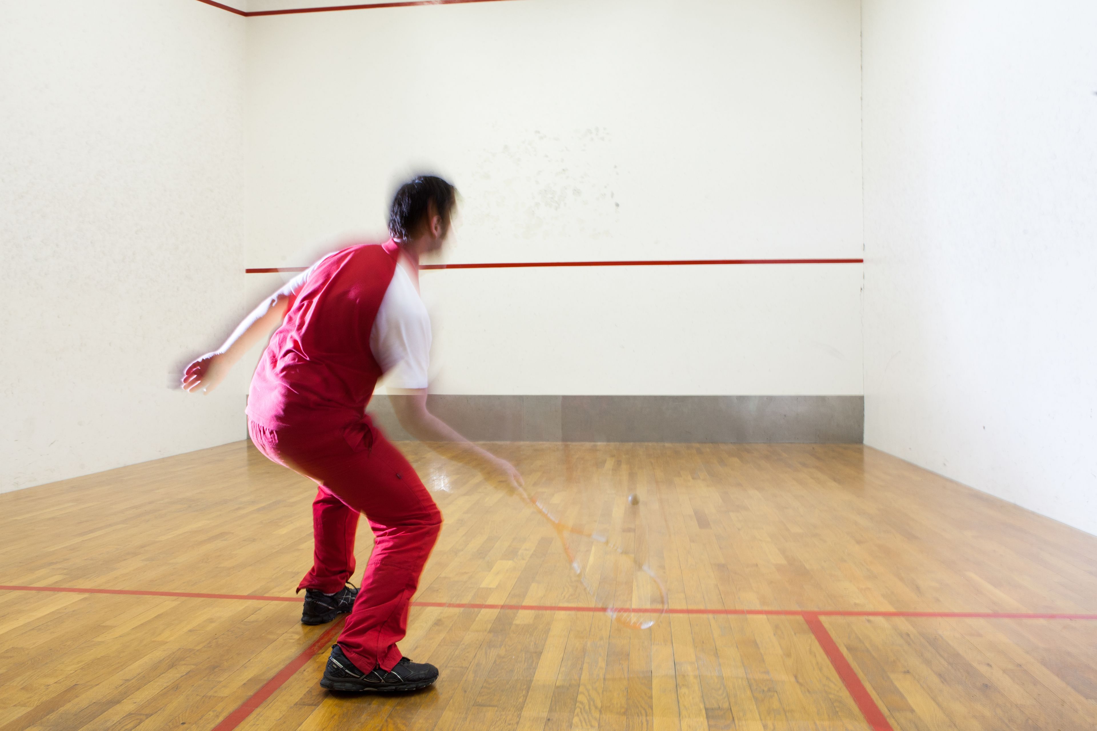 Eine Person spielt Squash in einem Raum mit Holzboden und roten Linien, die das Spielfeld markieren.