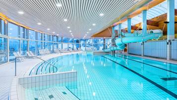 Una piscina interior con un tobogán azul, ventanas amplias y techo de madera.