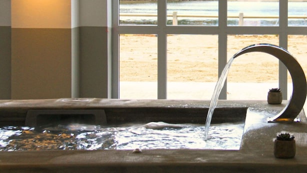 A water feature with a curved spout, a window with a view of a beach and a building, and a wall-mounted light fixture. - Hôtel du Parc (Soorts-Hossegor)