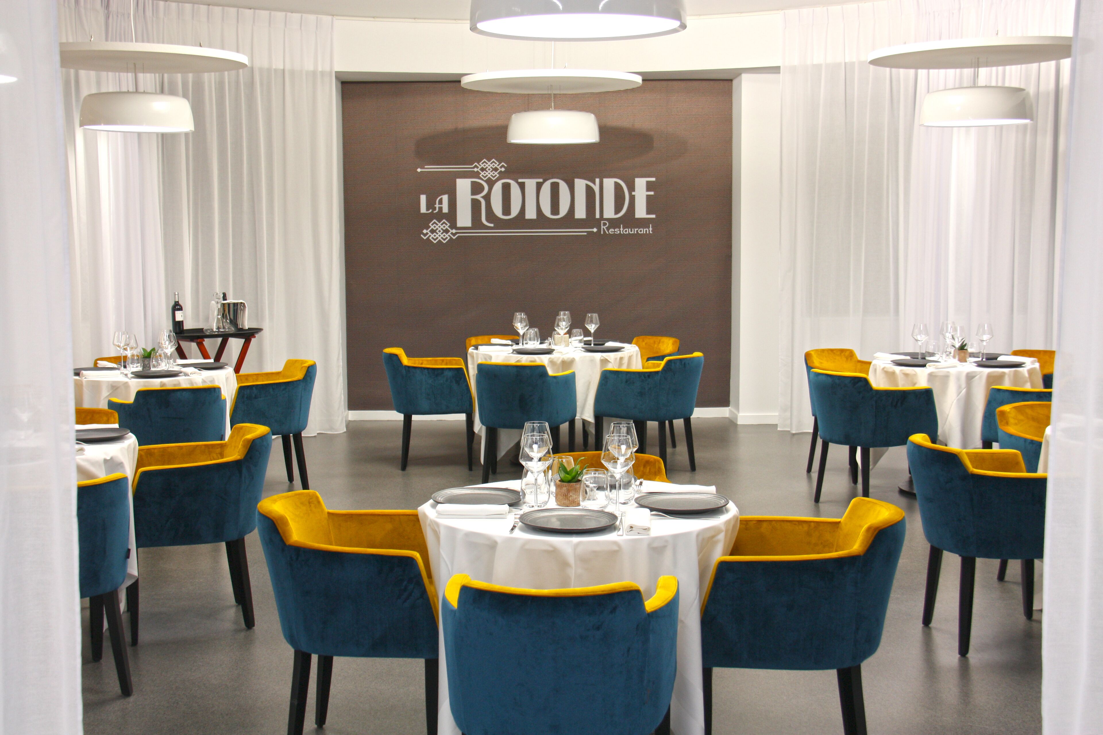 A restaurant interior with tables set for dining, blue and yellow chairs, and a sign reading "LA ROTONDE Restaurant".