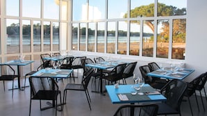 A restaurant with tables and chairs arranged for dining, large windows offering a view of the lake and trees, and a ceiling with skylights. - Hôtel du Parc (Soorts-Hossegor)