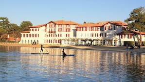 A large, multi-story building with a red-tiled roof, situated by a calm body of water. - Hôtel du Parc (Soorts-Hossegor)