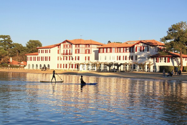 A large, multi-story building with a red-tiled roof, situated by a calm body of water.