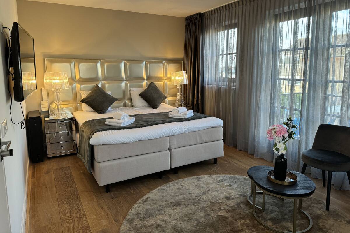 A modern hotel room with a large bed, a bedside table with lamps, a TV, a small round table with a vase, and a chair.