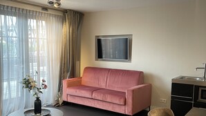 A modern living room with a pink sofa, a flat-screen TV mounted on the wall, a dining table with chairs, and a small round coffee table with a vase of flowers. - Najade Hotel (Loosdrecht)