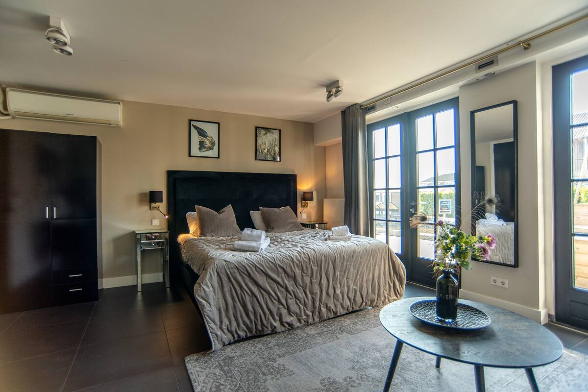 A modern hotel room with a large bed, a round table with a vase, and a view through the large windows.