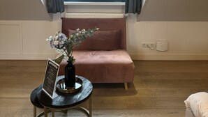 A room with a sloped ceiling, a pink sofa, a small table with a vase, and a window with curtains. - Najade Hotel (Loosdrecht)