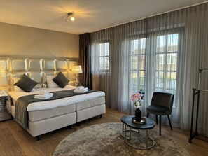 A hotel room with a large bed, a bedside table with a lamp, a chair, a small table with a vase, and large windows with curtains. - Najade Hotel (Loosdrecht)