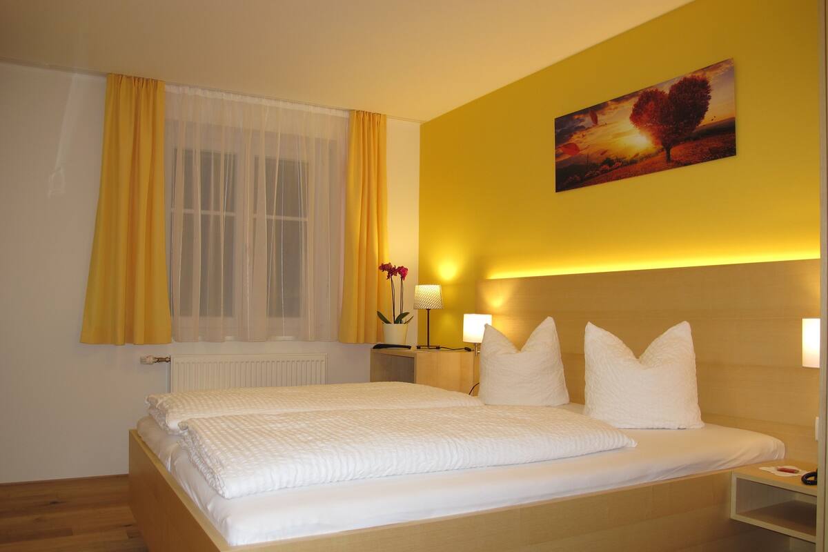 Een hotelkamer met een groot bed, een nachtkastje met een lamp, een raam met gele gordijnen en een schilderij aan de muur.