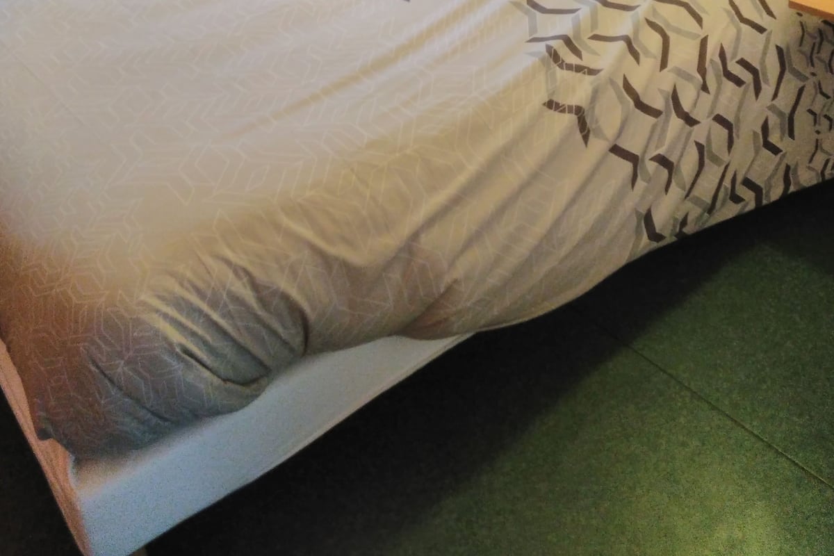 Un lit simple avec une couvre-lit à motifs, dans une chambre dotée d’une tête de lit en bois et d’une petite table en bois placée près de la fenêtre.