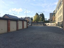 Un estacionamiento de grava con entrada protegida por un portón, edificios con fachada de ladrillo y revestimiento, y cielo despejado.