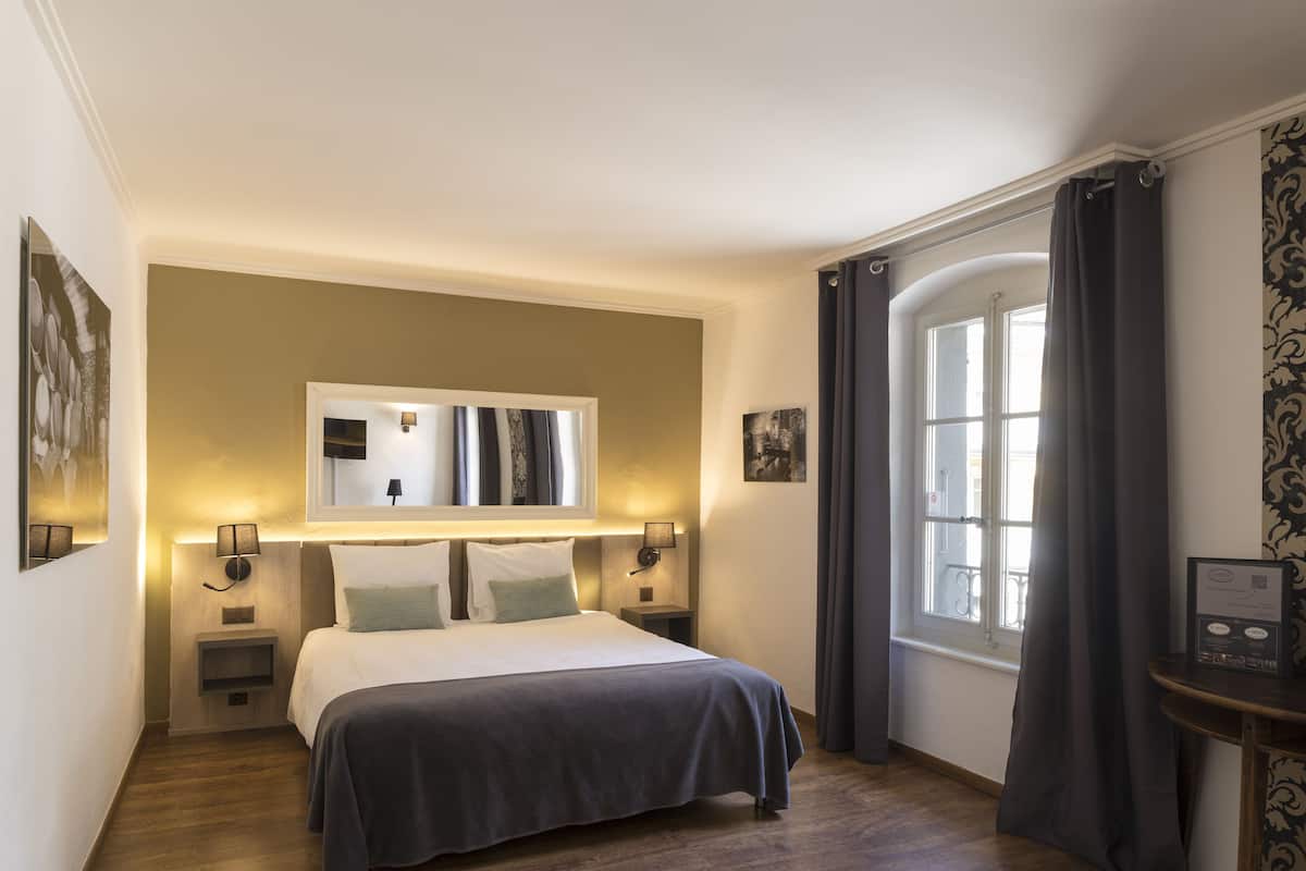 Habitación de hotel con cama, lámparas de noche, un espejo, una ventana con cortinas y una mesita con un menú.