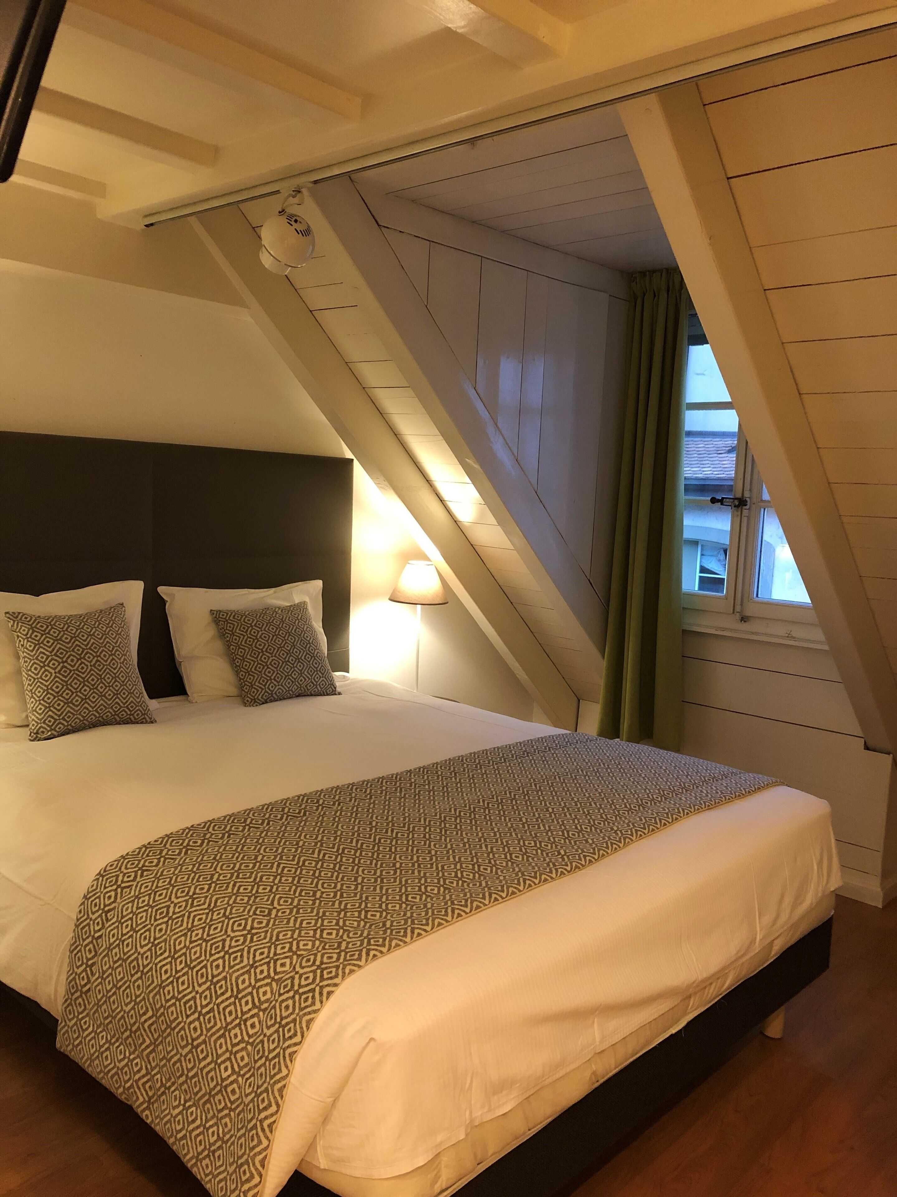 Une chambre à coucher avec un lit, des coussins à motifs, une lampe de chevet et une fenêtre avec des rideaux.