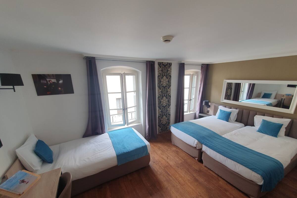 Habitación de hotel con dos camas individuales, suelo de madera, un ventanal grande con cortinas, un espejo montado en la pared y una mesita de noche pequeña.
