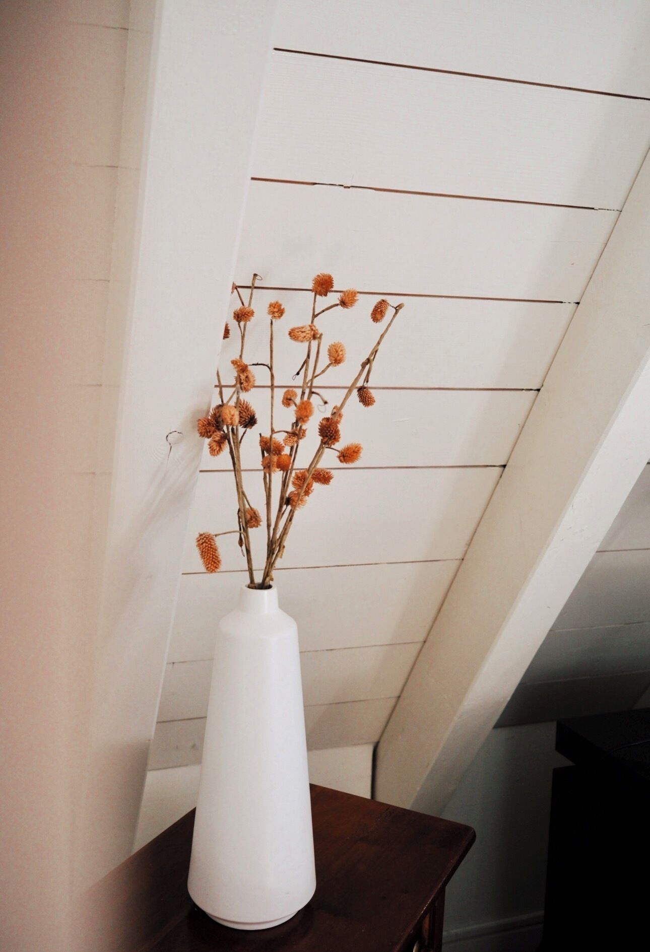 Un vase blanc contenant des fleurs séchées, posé sur une table en bois foncé, devant un mur blanc orné de lignes horizontales.