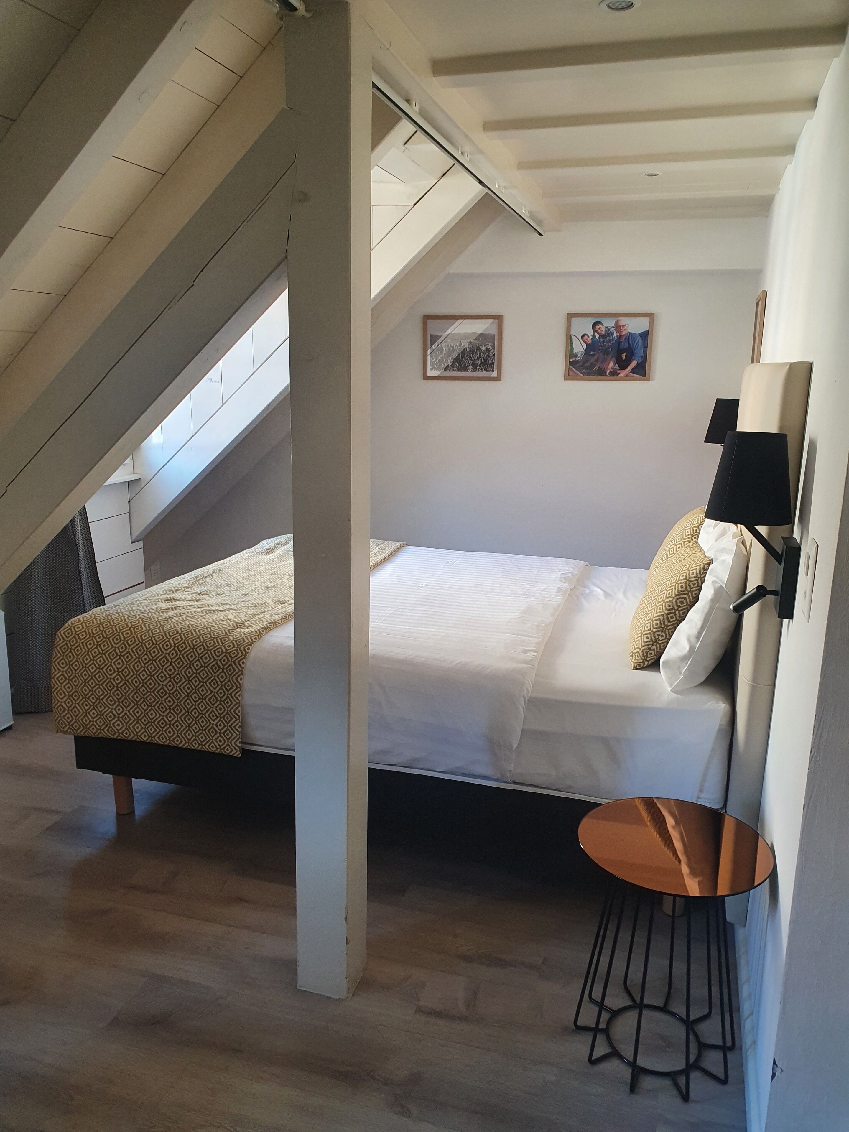 A bedroom with a sloped ceiling, a bed with a patterned bedspread, two pillows, a bedside lamp, and a small round table.