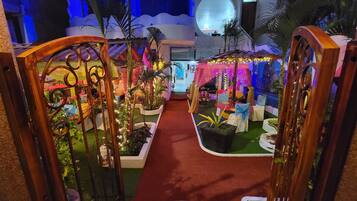 A tropical-themed indoor garden with a red carpet pathway, decorative plants, and a seating area with chairs and tables.
