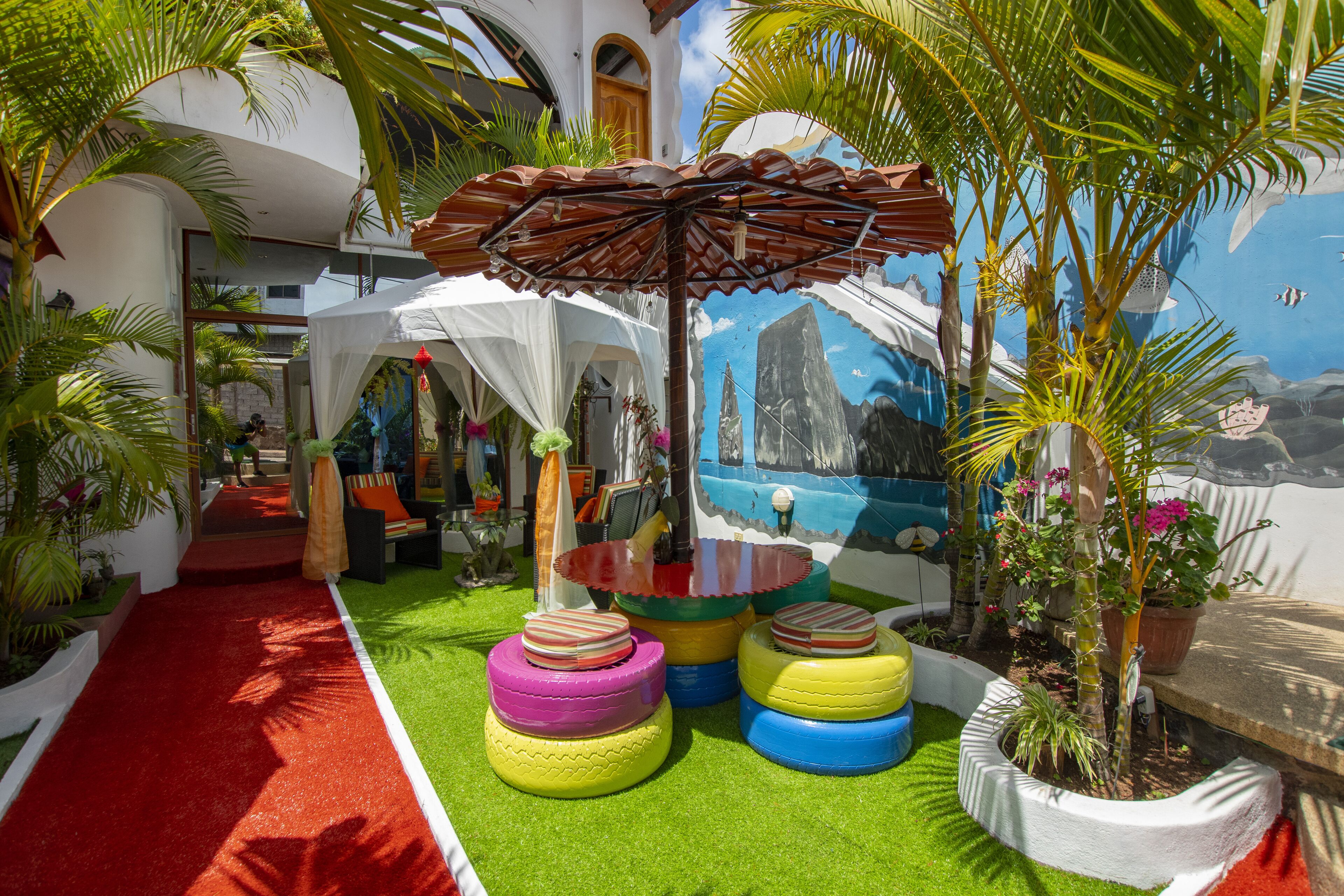 A tropical-themed outdoor area with a red carpet, colorful tire stools, a white tent, and a mural of a coastal scene.