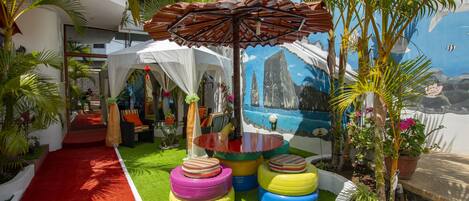 A tropical-themed outdoor area with a red carpet, colorful tire stools, a white tent, and a mural of a coastal scene.