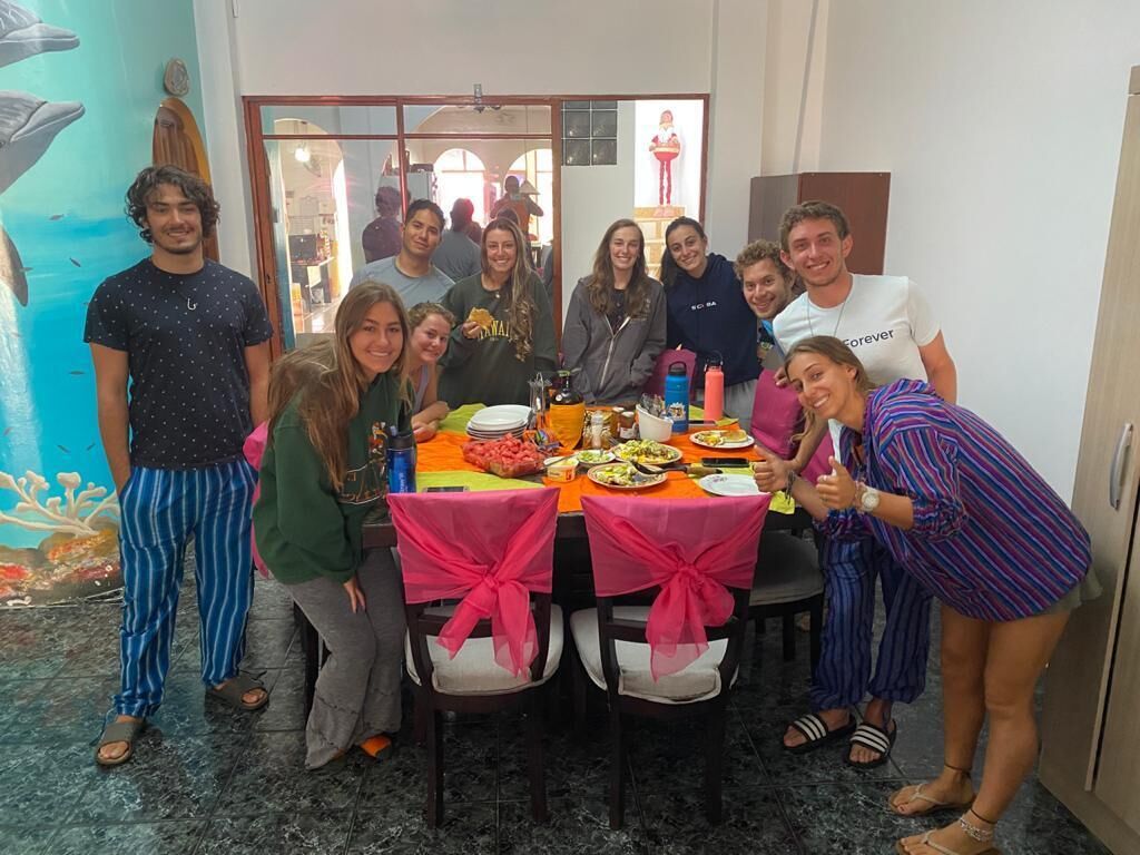 Eine Gruppe von Menschen hat sich um einen Tisch mit Essen und Getränken versammelt, in einem Raum mit Wandgemälde und einer Tür zu einem weiteren Bereich.
